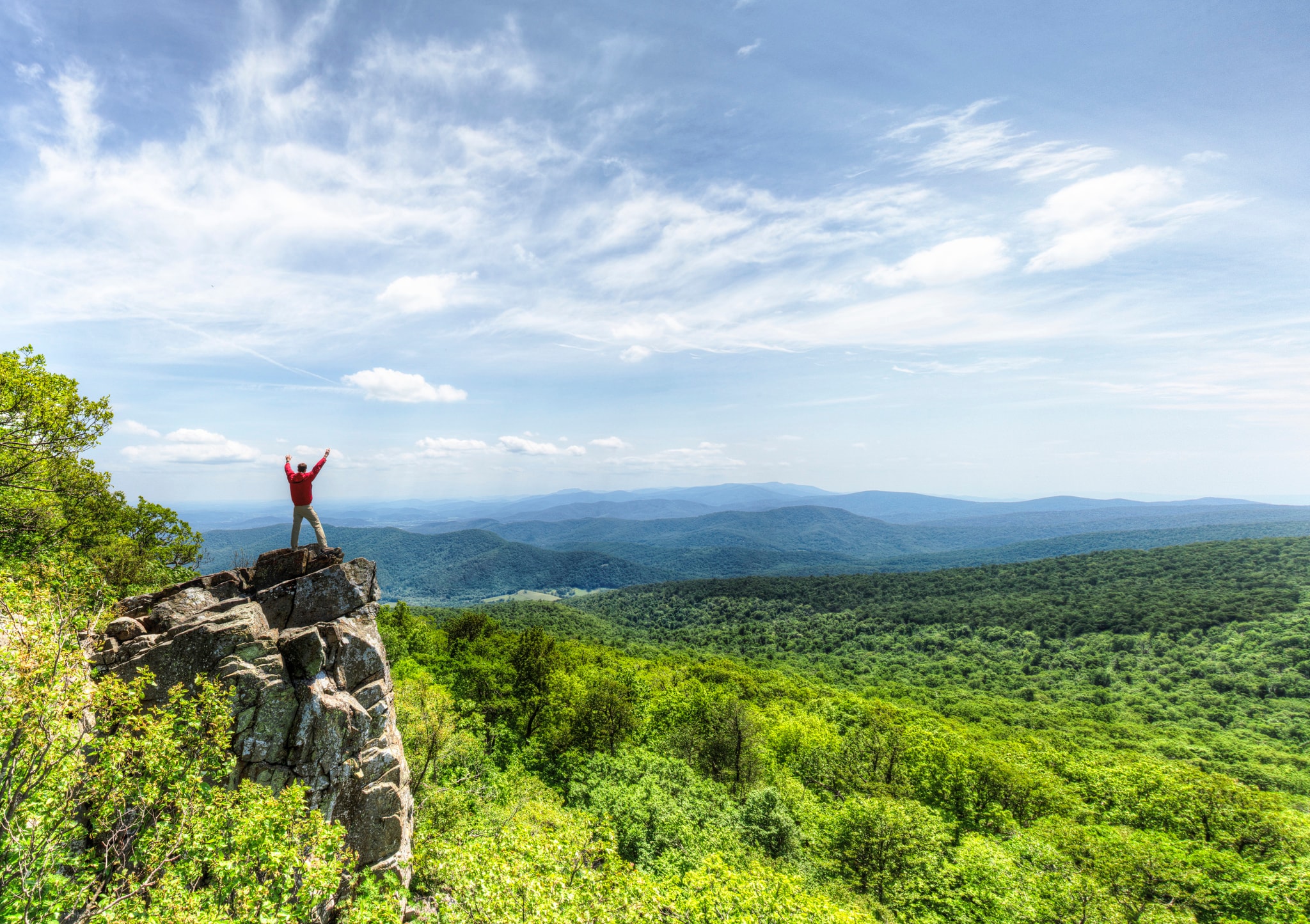 A man stands on a rocky outcrop overlooking the receding mountains.