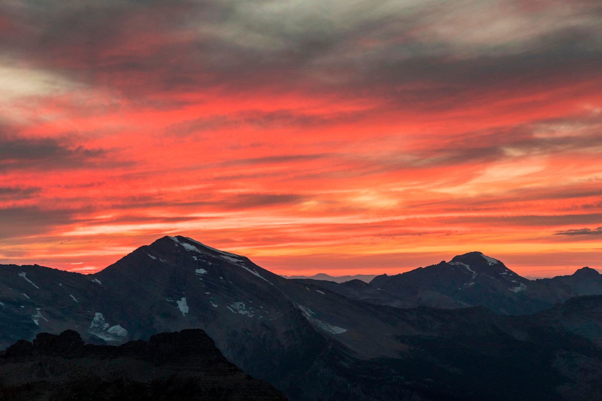 Clouds of orange and red sit above dark-gray mountains; snow dots the mountain peaks.